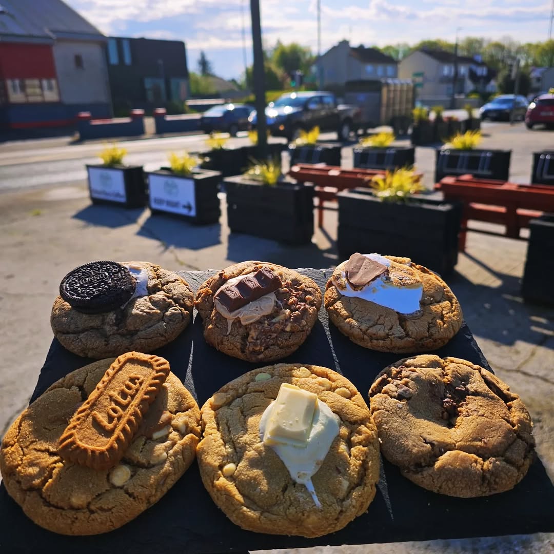 Six gourmet cookies with toppings displayed on a dark slate board outdoors.