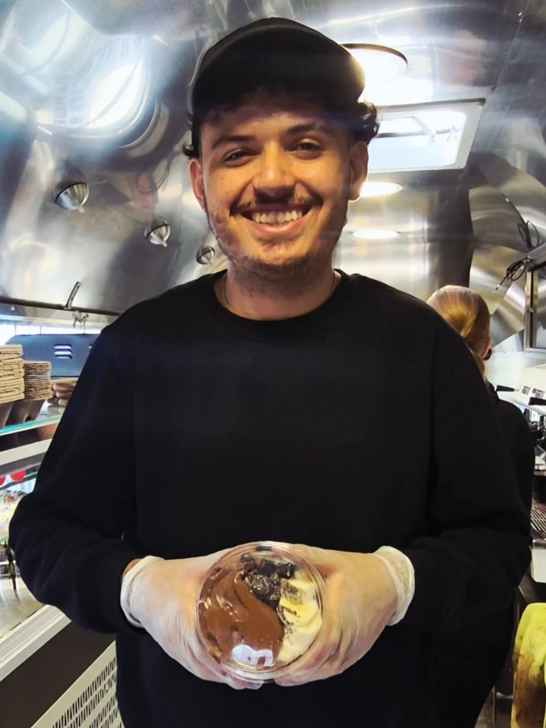 Smiling man in black shirt holding a layered dessert cup inside a food truck.